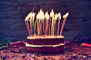 a cake topped with some lit candles before blowing out the cake, on a rustic wooden table full of confetti, party horns and streamers, with a filtered effect