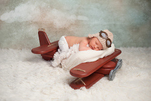 Studio portrait of an eight day old newborn baby boy wearing an aviator cap with goggles. He is sleeping on a vintage inspired airplane posing prop.