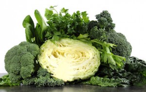 Healthy diet health foods with leafy green vegetables including cabbage, broccoli, broccolini, parsley, celery, silverbeet, and spinach on black slate kitchen top and white wall.